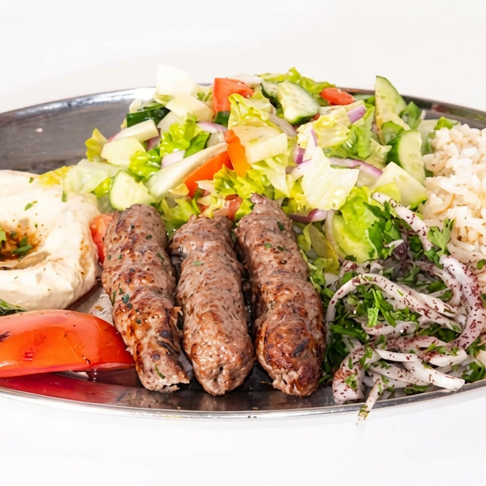 Grilled kebabs served with hummus, rice, chopped salad, sumac onions, and grilled peppers on a platter.